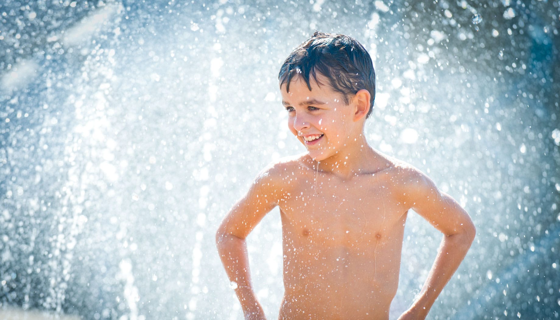kids-of-the-caribean A boy stands with hands on hips, smiling under a spray of water, with droplets glistening around him.